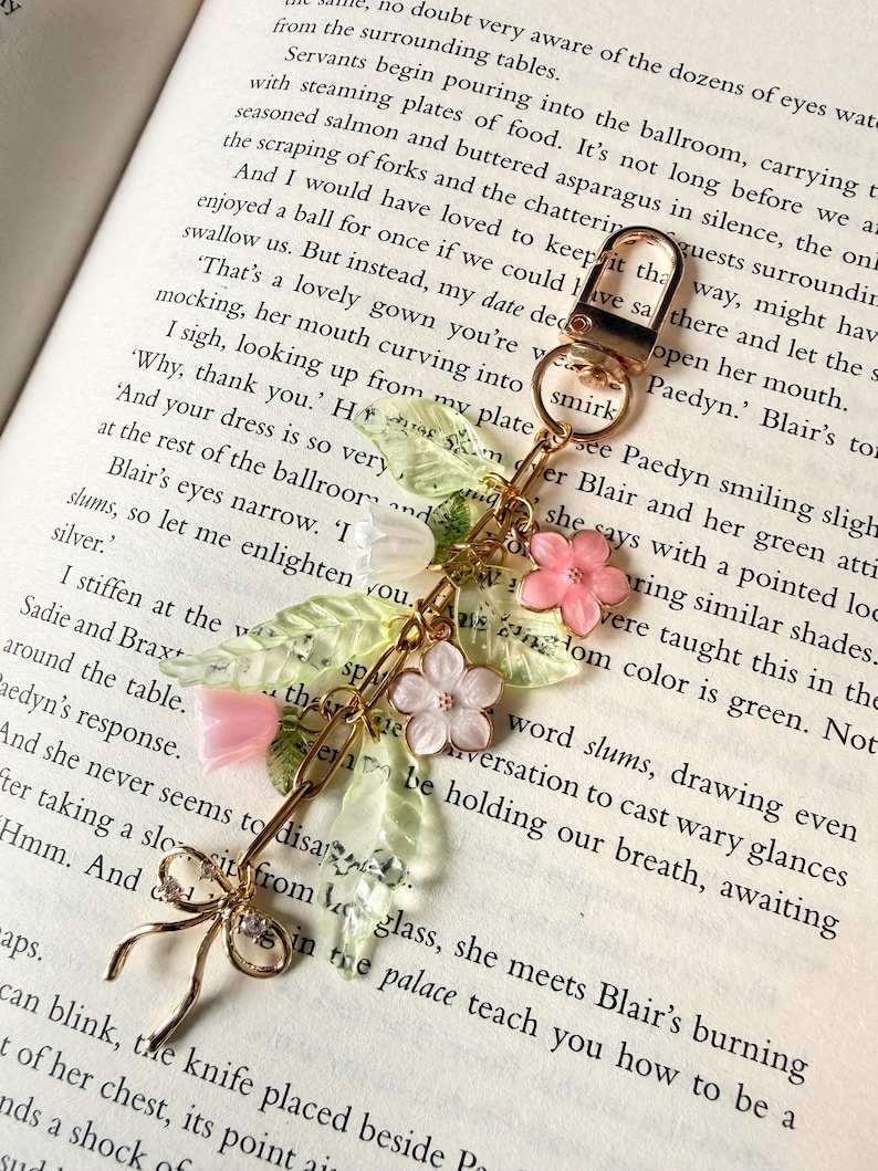 Puede incluir: Un llavero dorado con un dise&ntilde;o floral decorativo. El dije presenta flores rosas y blancas, hojas verdes y un lazo delicado. El llavero se muestra sobre el fondo de un libro abierto.