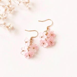 May include: A pair of drop earrings featuring clusters of small, translucent light pink flower-shaped charms. Each flower has a tiny gold-toned centre bead. The earrings are attached to gold-toned metal fishhook wires for pierced ears.