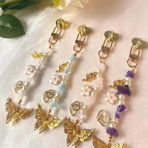 May include: Four decorative zipper pulls with gold-coloured hardware. Each pull features a gold butterfly charm, beads, and floral accents in various colours, including white, pink, blue, and purple. The pulls are designed for use on zippers.