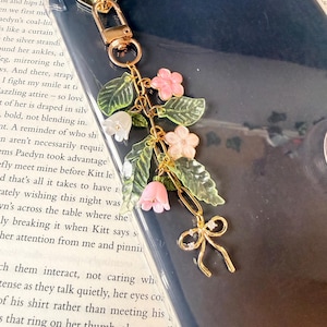 Puede incluir: Un charm de tel&eacute;fono dorado con un dise&ntilde;o floral. Presenta flores rosas, hojas verdes y un peque&ntilde;o lazo. El charm est&aacute; sujeto a un cierre dorado. El fondo es una funda de tel&eacute;fono azul oscuro.