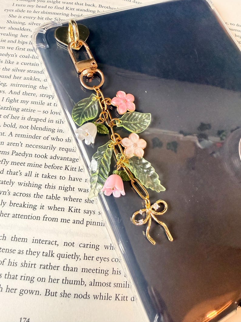 Puede incluir: Un charm de tel&eacute;fono dorado con un dise&ntilde;o floral. Presenta flores rosas, hojas verdes y un peque&ntilde;o lazo. El charm est&aacute; sujeto a un cierre dorado. El fondo es una funda de tel&eacute;fono azul oscuro.