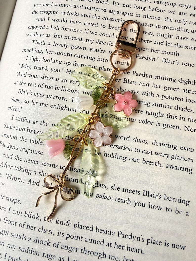 Puede incluir: Llavero dorado con dise&ntilde;o floral. El dije presenta flores rosas y blancas, hojas verdes y un lazo delicado. El llavero est&aacute; sobre el fondo de las p&aacute;ginas de un libro.