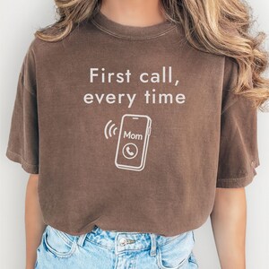 以下が含まれることがあります： 「First call, every time」の文字と電話のグラフィックがプリントされた茶色のTシャツ。電話のグラフィックは、画面に「Mom」という文字が表示された着信中の電話を示しています。カジュアルなスタイルのTシャツです。