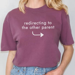 May include: A mauve-colored t-shirt with the text "redirecting to the other parent" and an arrow pointing right. The shirt is made of a soft material and has short sleeves. The model is wearing blue jeans.