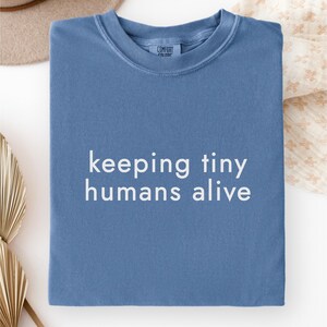 Può includere: Una t-shirt blu a girocollo con il testo bianco "keeping tiny humans alive" stampato in un semplice carattere sans-serif al centro. Il capo presenta un colletto a coste e un'etichetta del marchio visibile all'interno dello scollo.