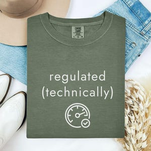 May include: An olive green t-shirt with the words "regulated (technically)" in white, along with a speedometer graphic. The shirt is laid flat with a hat, jeans, and white boots. The shirt is a Comfort Colors brand.
