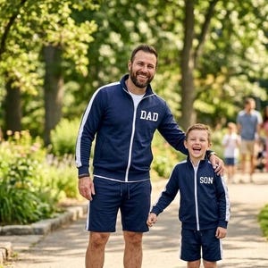 Può includere: Tute abbinate blu navy con strisce laterali bianche e cerniere bianche. La giacca da adulto presenta la scritta DAD in bianco, mentre la giacca da bambino presenta la scritta SON in bianco. Entrambi i set includono una giacca e pantaloncini.