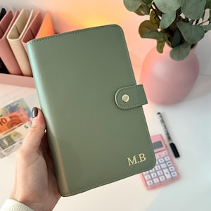 Puede incluir: Una agenda verde salvia con un cierre dorado y las iniciales "M.B." en letras doradas. La agenda se sostiene en una mano. Otros elementos en el fondo incluyen una calculadora rosa, un bolígrafo y un jarrón rosa con vegetación.