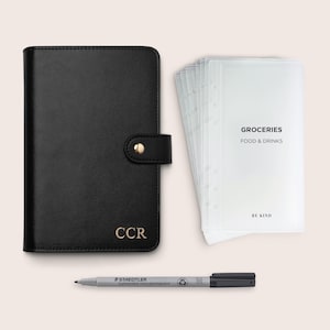 Op de afbeelding: Een zwarte leren planner met gouden initialen en een drukknoopsluiting. Inclusief transparante plastic inzetstukken met de tekst "GROCERIES FOOD & DRINKS" en een zwarte marker. De tekst "BE KIND" is ook zichtbaar.