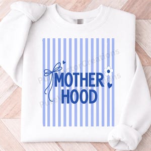 Peut inclure: Sweat-shirt blanc avec des rayures verticales bleues et le mot "MOTHERHOOD" en lettres bleues. Un nœud bleu, une fleur et des cœurs ornent le design. Le sweat-shirt a des manches longues et un col rond.