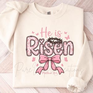 May include: Cream-colored sweatshirt with the words "He is Risen" in pink, floral-patterned letters. A pink bow and butterflies accent the design, with a crown of thorns above the word "Risen". The text "Matthew 28:6" is below the bow.
