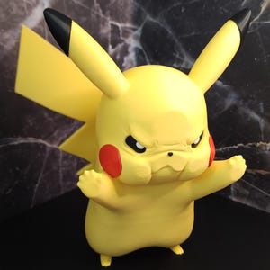 May include: A yellow Pikachu figurine with a stern expression, red cheek circles, and black-tipped ears. The figure has its arms out and is set against a dark background.