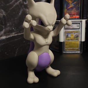 May include: A light grey 3D-printed Mewtwo figure with purple detailing, posed with raised arms. The background features graded Pokémon trading cards. The figure is a collectible item.