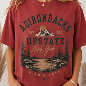 K&ouml;nnte beinhalten: Ein ziegelrotes Kurzarm-T-Shirt mit einem dunkelgr&uuml;nen Grafikdruck von Bergen, einem See und Tannen. Der Text lautet ADIRONDACKS, EST. 1892, UPSTATE, New York und WILD & FREE. Das Design enth&auml;lt eine Sonne und kleine V&ouml;gel.