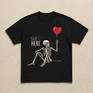 Pu&ograve; includere: Una t-shirt nera a maniche corte con la grafica di uno scheletro seduto che tiene in mano un palloncino rosso a forma di cuore. Il testo recita Still HERE con una piccola icona a forma di cuore e la frase That's growth. in inchiostro bianco e rosso.