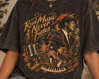 Könnte beinhalten: Ein anthrazitfarbenes Kurzarm-T-Shirt mit einem Grafikdruck von zwei Jazzmusikern mit Hüten, Mikrofonen, einer Schallplatte und Klaviertasten. Der Text lautet Real Music Never Fades in einer stilisierten goldenen Schrift mit einer kleinen Krone.