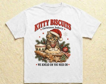 Könnte beinhalten: Ein weißes T-Shirt mit einer Grafik einer getigerten Katze, die eine rote Weihnachtsmütze und eine Schürze trägt und Teig auf einem Holzbrett knetet. Der Text lautet KITTY BISCUITS christmas bakery und WE KNEAD EM YOU NEED EM.