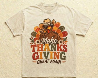Könnte beinhalten: Ein beiges Kurzarm-T-Shirt mit einer Grafik eines Truthahns, der einen Cowboyhut und eine Sonnenbrille trägt und ein Friedenszeichen macht. Der Text lautet Make THANKS GIVING GREAT AGAIN, umgeben von bunten Federn und Sonnenblumen.