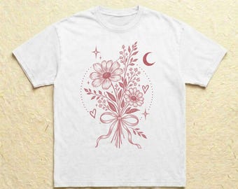 Könnte beinhalten: Ein weißes T-Shirt mit kurzen Ärmeln und einem mittigen Aufdruck in Altrosa. Das Motiv zeigt einen Blumenstrauß, der mit einer Schleife gebunden ist, umgeben von kleinen Sternen, einer Mondsichel und Herzen in einem gepunkteten Kreis.