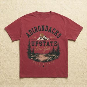 K&ouml;nnte beinhalten: Ein verwaschenes rotes Kurzarm-T-Shirt mit einem Grafikdruck einer Berglandschaft mit See und Tannen. Der Text ADIRONDACKS EST. 1892 UPSTATE New York WILD & FREE ist in dunkelgr&uuml;ner und beiger Schrift mittig auf der Brust abgebildet.
