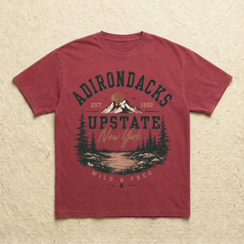 K&ouml;nnte beinhalten: Ein verwaschenes rotes Kurzarm-T-Shirt mit einem Grafikdruck einer Berglandschaft mit See und Tannen. Der Text ADIRONDACKS EST. 1892 UPSTATE New York WILD & FREE ist in dunkelgr&uuml;ner und beiger Schrift mittig auf der Brust abgebildet.