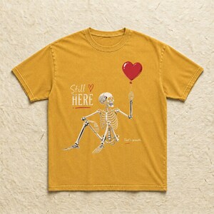 Pu&ograve; includere: Una t-shirt giallo senape con la grafica di uno scheletro seduto che tiene in mano un palloncino rosso a forma di cuore. Il testo "Still HERE" &egrave; stampato in bianco sopra lo scheletro, con un piccolo cuore rosso e "That's growth" sotto.