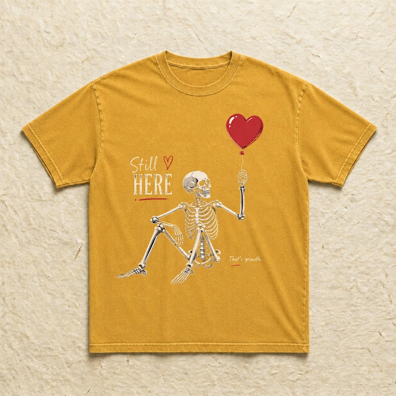 Pu&ograve; includere: Una t-shirt giallo senape con la grafica di uno scheletro seduto che tiene in mano un palloncino rosso a forma di cuore. Il testo "Still HERE" &egrave; stampato in bianco sopra lo scheletro, con un piccolo cuore rosso e "That's growth" sotto.