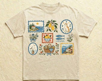 Könnte beinhalten: Ein beiges T-Shirt mit kurzen Ärmeln und einer Ansammlung bunter Illustrationen, darunter eine Küstenlandschaft, ein Zitronenzweig, eine sitzende Katze, eine Möwe, ein Blumenkorb, zwei Fische und ein mediterranes Dorf mit einem Segelboot.