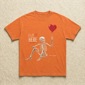 Pu&ograve; includere: Una t-shirt arancione a maniche corte con la grafica di uno scheletro seduto che tiene in mano un palloncino rosso a forma di cuore. Il testo "Still HERE" &egrave; stampato in bianco sopra lo scheletro e le parole "That's growth." appaiono sotto.
