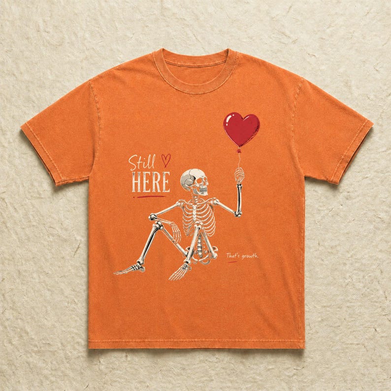 Pu&ograve; includere: Una t-shirt arancione a maniche corte con la grafica di uno scheletro seduto che tiene in mano un palloncino rosso a forma di cuore. Il testo "Still HERE" &egrave; stampato in bianco sopra lo scheletro e le parole "That's growth." appaiono sotto.