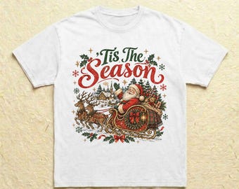 Könnte beinhalten: Ein weißes T-Shirt mit einem Grafikdruck von Santa Claus in einem von zwei Rentieren gezogenen Schlitten. Der Text 'Tis The Season' ist in Rot und Grün über der Illustration gedruckt, die von kleinen roten und grünen Schneeflocken umgeben ist.