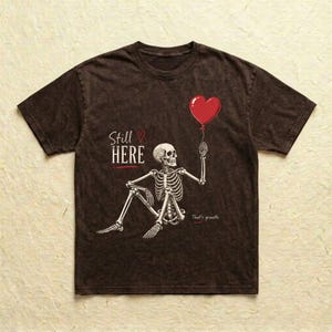 Pu&ograve; includere: Una t-shirt marrone scuro con effetto slavato che presenta la grafica di uno scheletro seduto che tiene in mano un palloncino rosso a forma di cuore. Il testo Still HERE con un piccolo cuore rosso e la scritta That's growth sono in bianco.