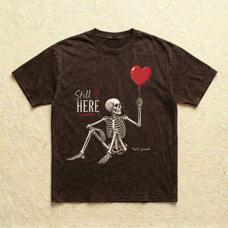 Pu&ograve; includere: Una t-shirt marrone scuro con effetto slavato che presenta la grafica di uno scheletro seduto che tiene in mano un palloncino rosso a forma di cuore. Il testo Still HERE con un piccolo cuore rosso e la scritta That's growth sono in bianco.