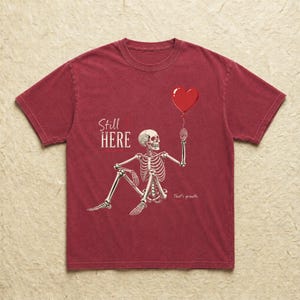 Pu&ograve; includere: Una t-shirt a maniche corte rosso scuro con la grafica di uno scheletro bianco seduto che tiene in mano un palloncino rosso a forma di cuore. Il testo "Still HERE" &egrave; stampato in bianco sopra lo scheletro e "That's growth." sotto.