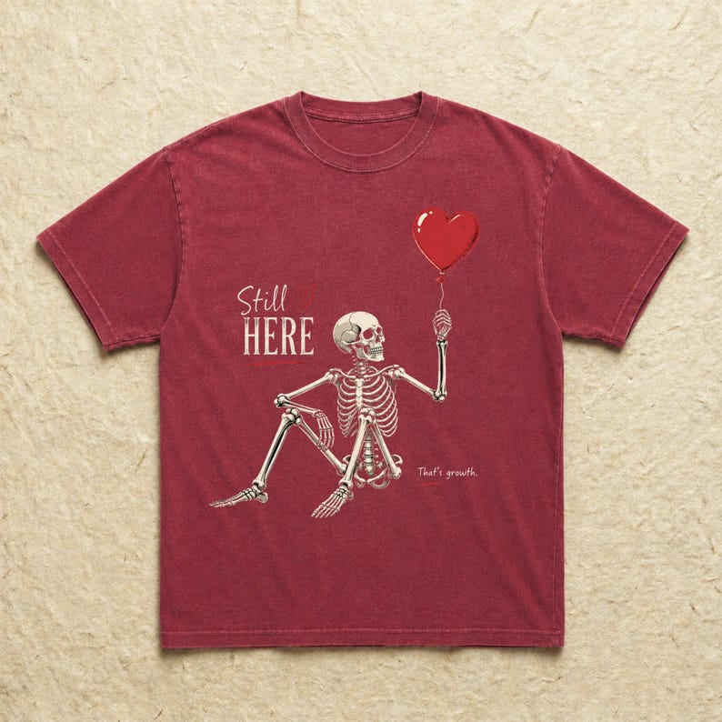 Pu&ograve; includere: Una t-shirt a maniche corte rosso scuro con la grafica di uno scheletro bianco seduto che tiene in mano un palloncino rosso a forma di cuore. Il testo "Still HERE" &egrave; stampato in bianco sopra lo scheletro e "That's growth." sotto.