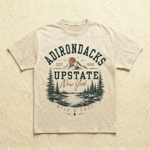 K&ouml;nnte beinhalten: Ein beiges Kurzarm-T-Shirt mit einer dunkelgr&uuml;nen und braunen Grafik einer Berglandschaft mit Kiefern, einem See und dem Text ADIRONDACKS EST. 1892 UPSTATE New York WILD & FREE, der kreisf&ouml;rmig auf der Vorderseite aufgedruckt ist.