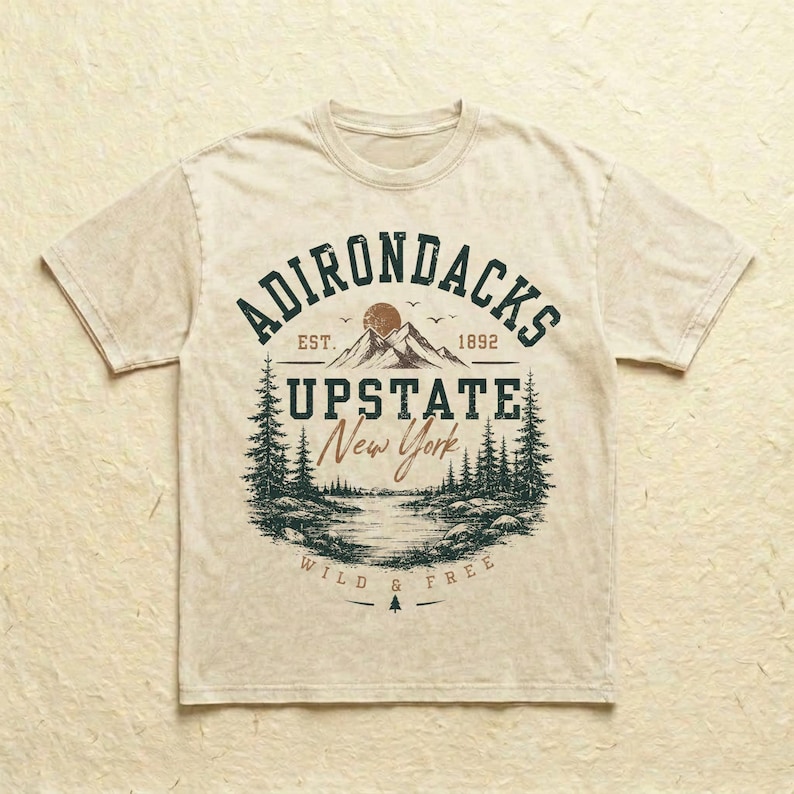 K&ouml;nnte beinhalten: Ein beiges Kurzarm-T-Shirt mit einer dunkelgr&uuml;nen und braunen Grafik einer Berglandschaft mit Kiefern, einem See und dem Text ADIRONDACKS EST. 1892 UPSTATE New York WILD & FREE, der kreisf&ouml;rmig auf der Vorderseite aufgedruckt ist.