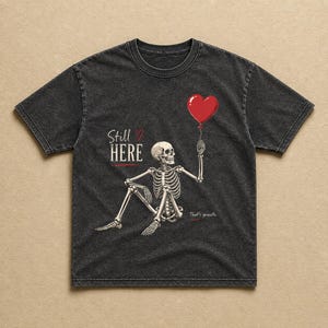 Pu&ograve; includere: Una t-shirt grigio antracite effetto slavato con la grafica di uno scheletro seduto che tiene in mano un palloncino rosso a forma di cuore. Il testo recita Still HERE con un piccolo cuore e That's growth. Il disegno &egrave; stampato in bianco e rosso.