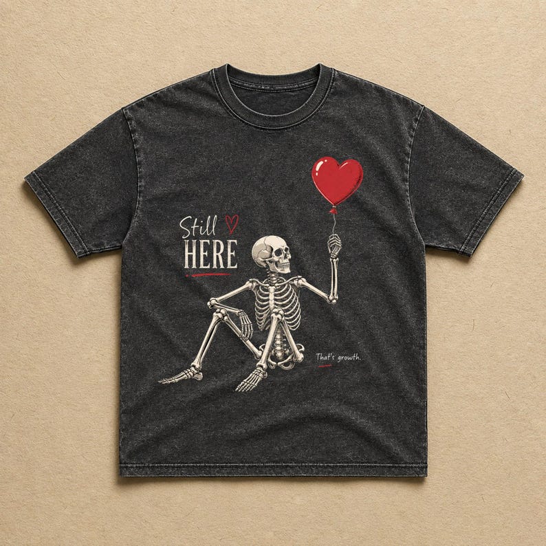 Pu&ograve; includere: Una t-shirt grigio antracite effetto slavato con la grafica di uno scheletro seduto che tiene in mano un palloncino rosso a forma di cuore. Il testo recita Still HERE con un piccolo cuore e That's growth. Il disegno &egrave; stampato in bianco e rosso.