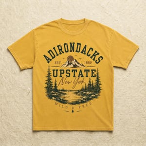 K&ouml;nnte beinhalten: Ein senfgelbes Kurzarm-T-Shirt mit einem dunkelgr&uuml;nen Grafikdruck einer Berglandschaft mit Tannen, einem See und dem Text ADIRONDACKS EST. 1892 UPSTATE New York WILD & FREE, der mittig auf der Vorderseite des Kleidungsst&uuml;cks platziert ist.