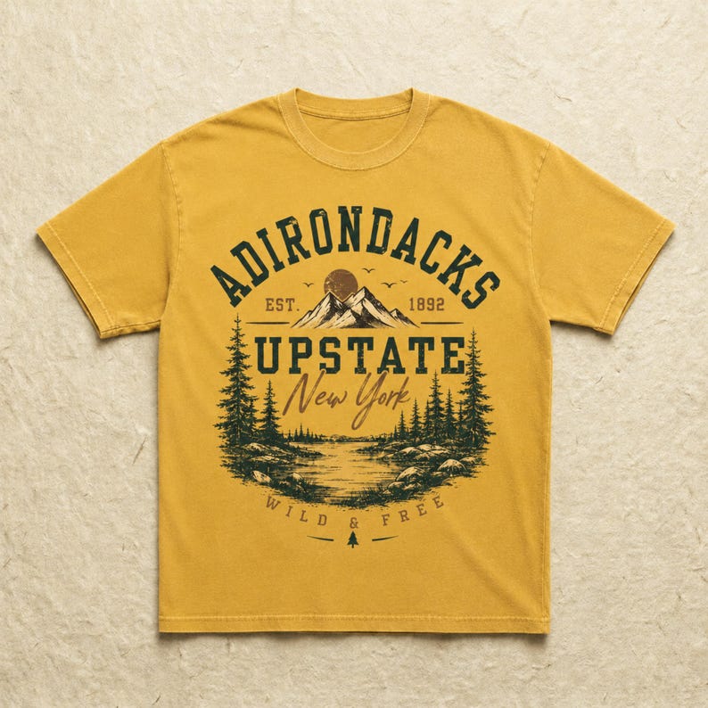 K&ouml;nnte beinhalten: Ein senfgelbes Kurzarm-T-Shirt mit einem dunkelgr&uuml;nen Grafikdruck einer Berglandschaft mit Tannen, einem See und dem Text ADIRONDACKS EST. 1892 UPSTATE New York WILD & FREE, der mittig auf der Vorderseite des Kleidungsst&uuml;cks platziert ist.