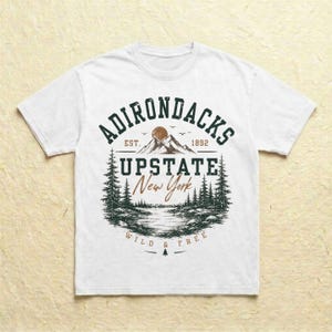 K&ouml;nnte beinhalten: Ein wei&szlig;es T-Shirt mit einem dunkelgr&uuml;nen und braunen Grafikdruck. Das Design enth&auml;lt den Text ADIRONDACKS EST. 1892, UPSTATE New York und WILD & FREE sowie eine Illustration von Bergen, einer Sonne, Tannen und einem See.