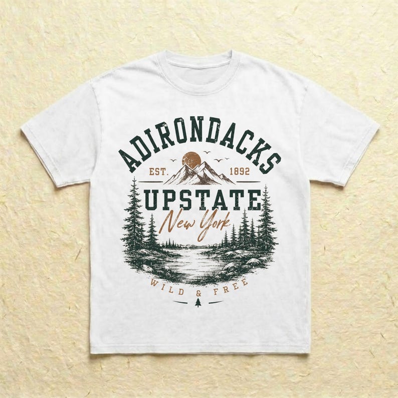 K&ouml;nnte beinhalten: Ein wei&szlig;es T-Shirt mit einem dunkelgr&uuml;nen und braunen Grafikdruck. Das Design enth&auml;lt den Text ADIRONDACKS EST. 1892, UPSTATE New York und WILD & FREE sowie eine Illustration von Bergen, einer Sonne, Tannen und einem See.