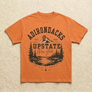 K&ouml;nnte beinhalten: Ein orangefarbenes T-Shirt mit kurzen &Auml;rmeln und einem dunklen Grafikdruck einer Berglandschaft mit Kiefern, einem See und einer Sonne. Der Text lautet ADIRONDACKS EST. 1892 UPSTATE New York WILD & FREE, angeordnet in einem zentrierten Design.