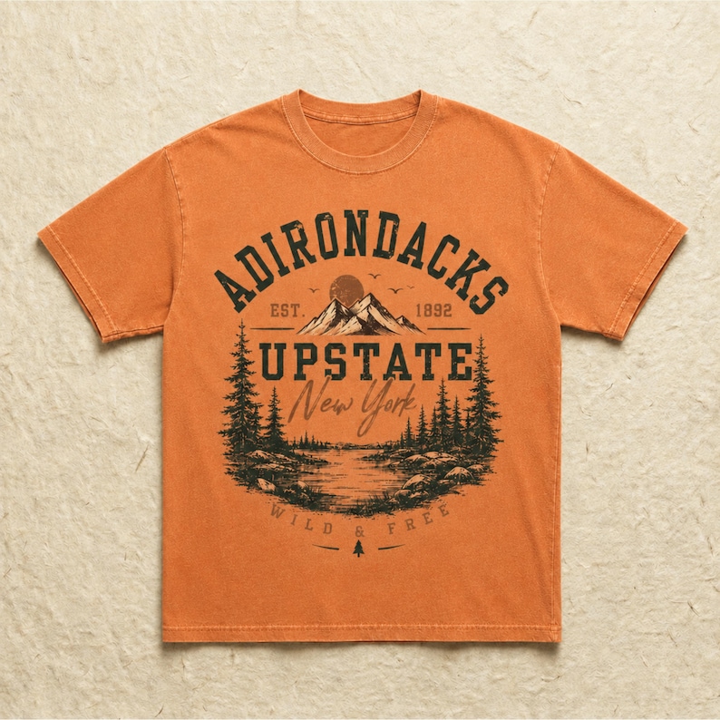 K&ouml;nnte beinhalten: Ein orangefarbenes T-Shirt mit kurzen &Auml;rmeln und einem dunklen Grafikdruck einer Berglandschaft mit Kiefern, einem See und einer Sonne. Der Text lautet ADIRONDACKS EST. 1892 UPSTATE New York WILD & FREE, angeordnet in einem zentrierten Design.