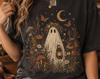 Könnte beinhalten: Ein anthrazitfarbenes Kurzarm-T-Shirt mit einer Grafik eines Geistes, der einen Blumenkranz trägt und eine Laterne hält, umgeben von Wildblumen, Schmetterlingen und einer Mondsichel. Das Shirt hat ein Comfort Colors-Etikett am Halsausschnitt.