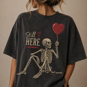 Pu&ograve; includere: Una t-shirt grigio antracite con la grafica di uno scheletro seduto che tiene in mano un palloncino rosso a forma di cuore. Il testo recita Still HERE con una piccola icona a forma di cuore e la frase That's growth. Design dall'aspetto vintage.