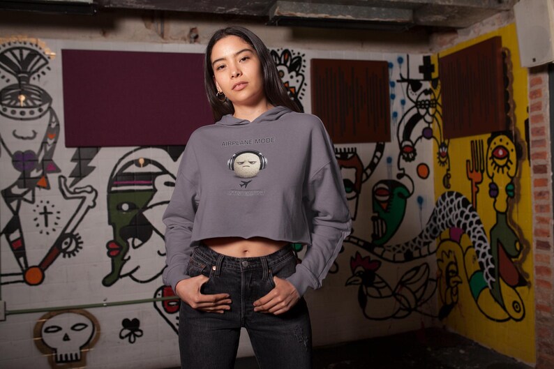 May include: A gray cropped hoodie with the text "PRIVATE MODE" and a graphic of a face wearing headphones. The person is wearing black jeans. The background is a graffiti-covered wall.