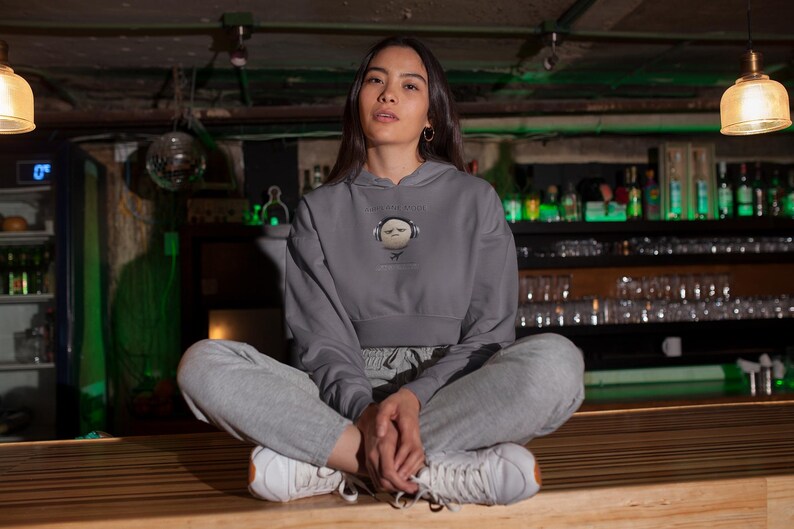 May include: A person wearing a gray cropped hoodie with a graphic of a character wearing headphones. The person is also wearing gray sweatpants and white sneakers. The background includes a bar setting with bottles and glasses.