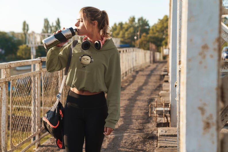 May include: An olive green cropped hoodie with a graphic of a face wearing headphones. The person is drinking from a silver and black water bottle. Black leggings and a black and red gym bag complete the athletic look.